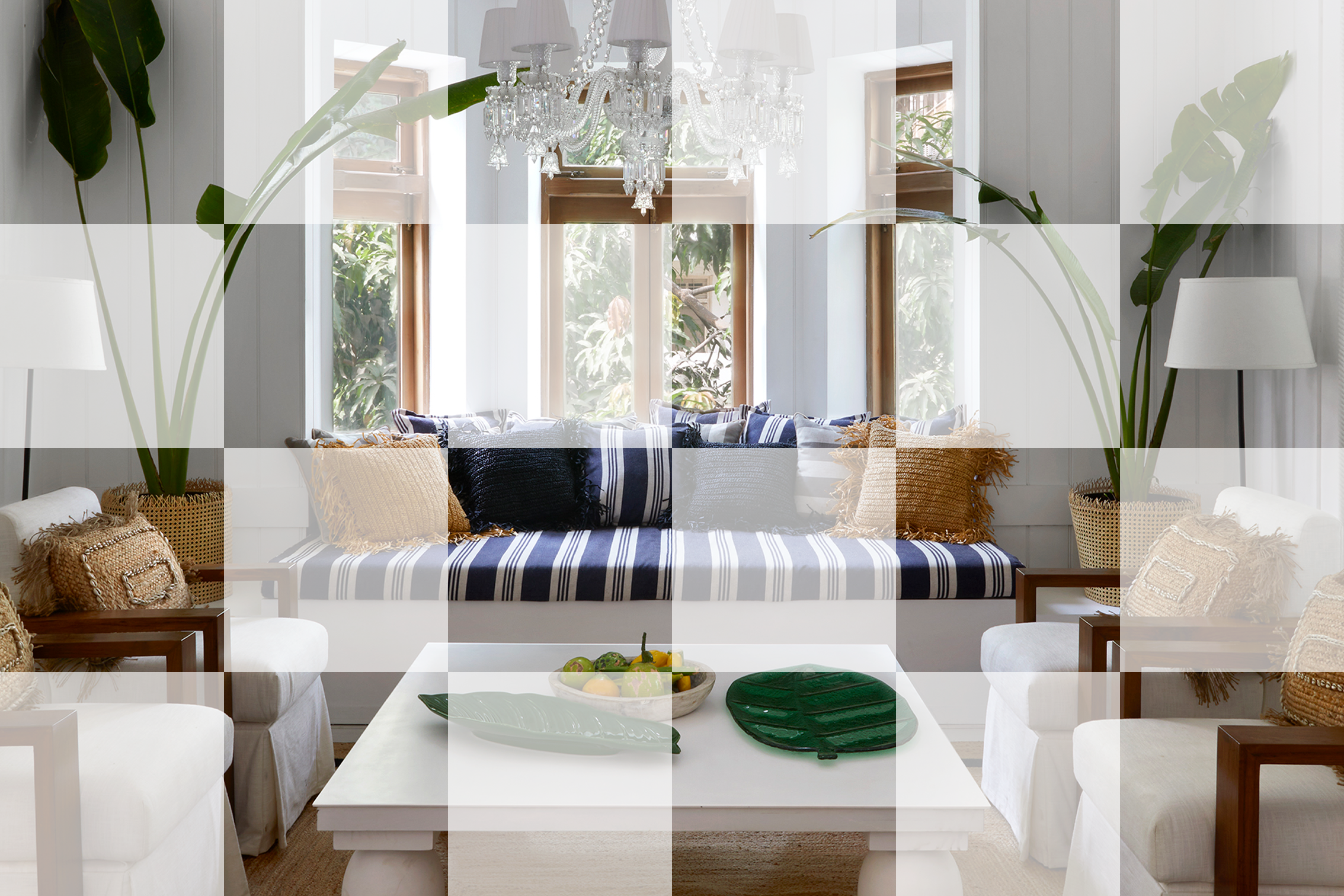 Living room with white panel walls, large windows, and a white chandelier. Contains a white sofa with striped and dark blue pillows, two white armchairs with beige pillows and wooden armrests, a white coffee table with decorative green dishes and a fruit bowl, and tall green plants flanking the sofa.