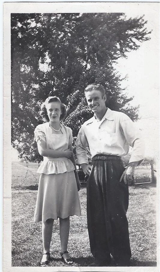 A black-and-white photo of a young woman and man standing outdoors in front of a large tree. The woman is wearing a dress with puffed sleeves and a ruffled hem, and the man is wearing a button-up shirt and high-waisted pants. They are smiling and posing for the picture.