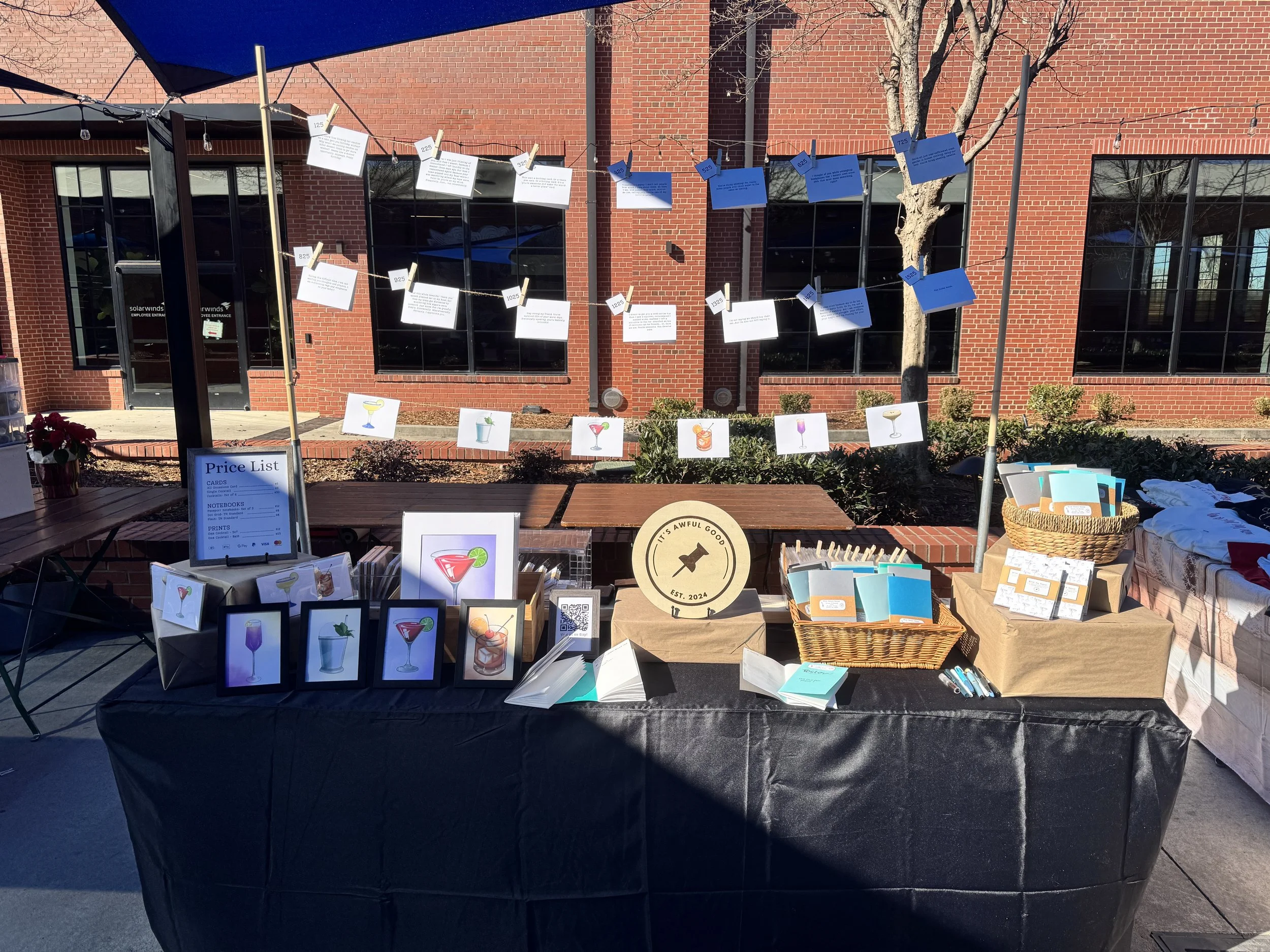 Outdoor booth with various printed products such as images of cocktails, notebooks, and price list. Decorations include hanging paper cards with descriptions, a circular sign that reads 'It’s Awful Good,' and other small items on tables. A large umbrella provides shade, and the background features a brick building with windows and a leafless tree.