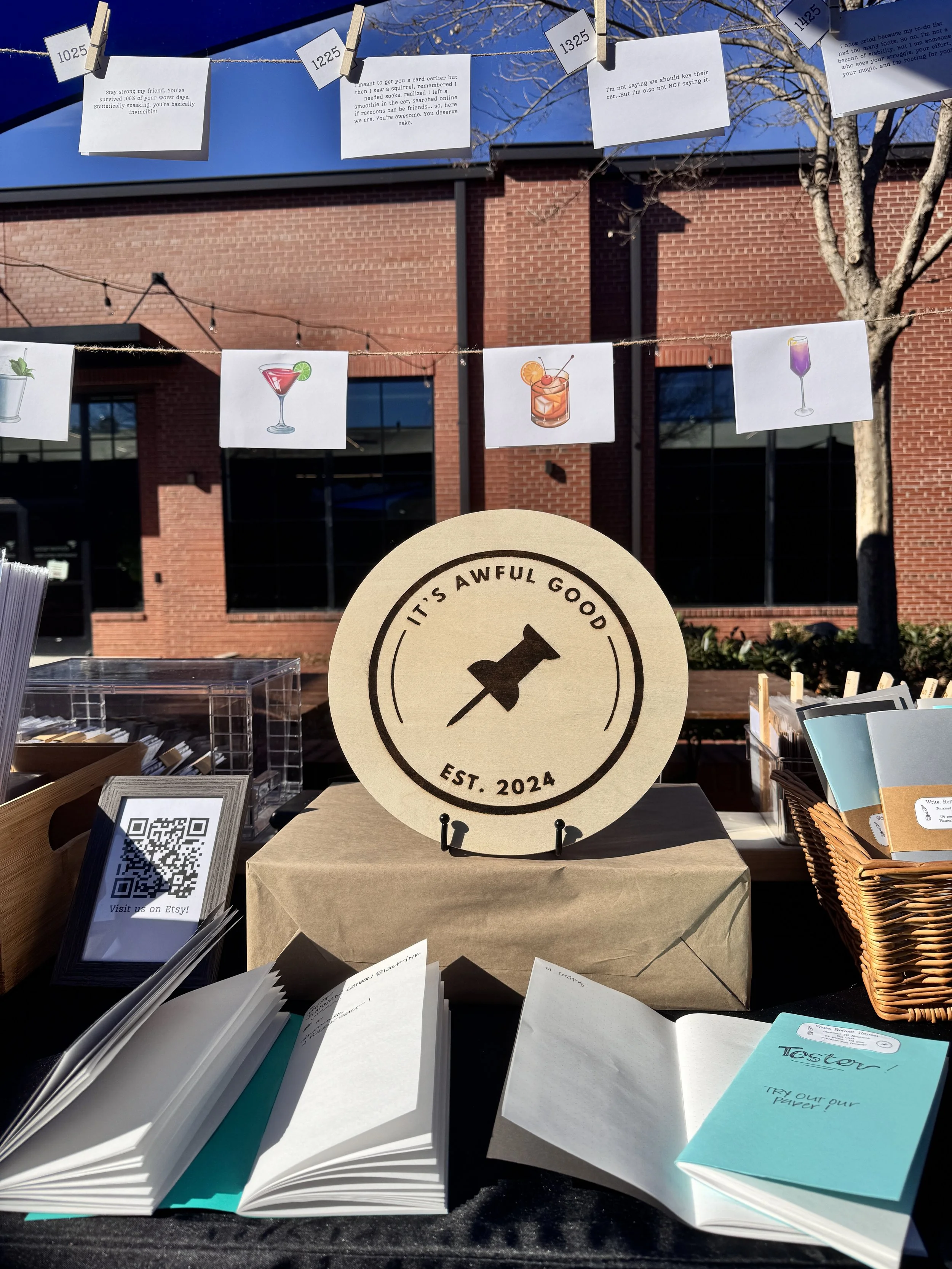 A display table with an emblem that reads 'IT'S AWFUL GOOD, EST. 2024' featuring a pushpin icon. Surrounding the table are bookmarks and booklets, with decorative banners hanging above showing illustrations of various cocktails.