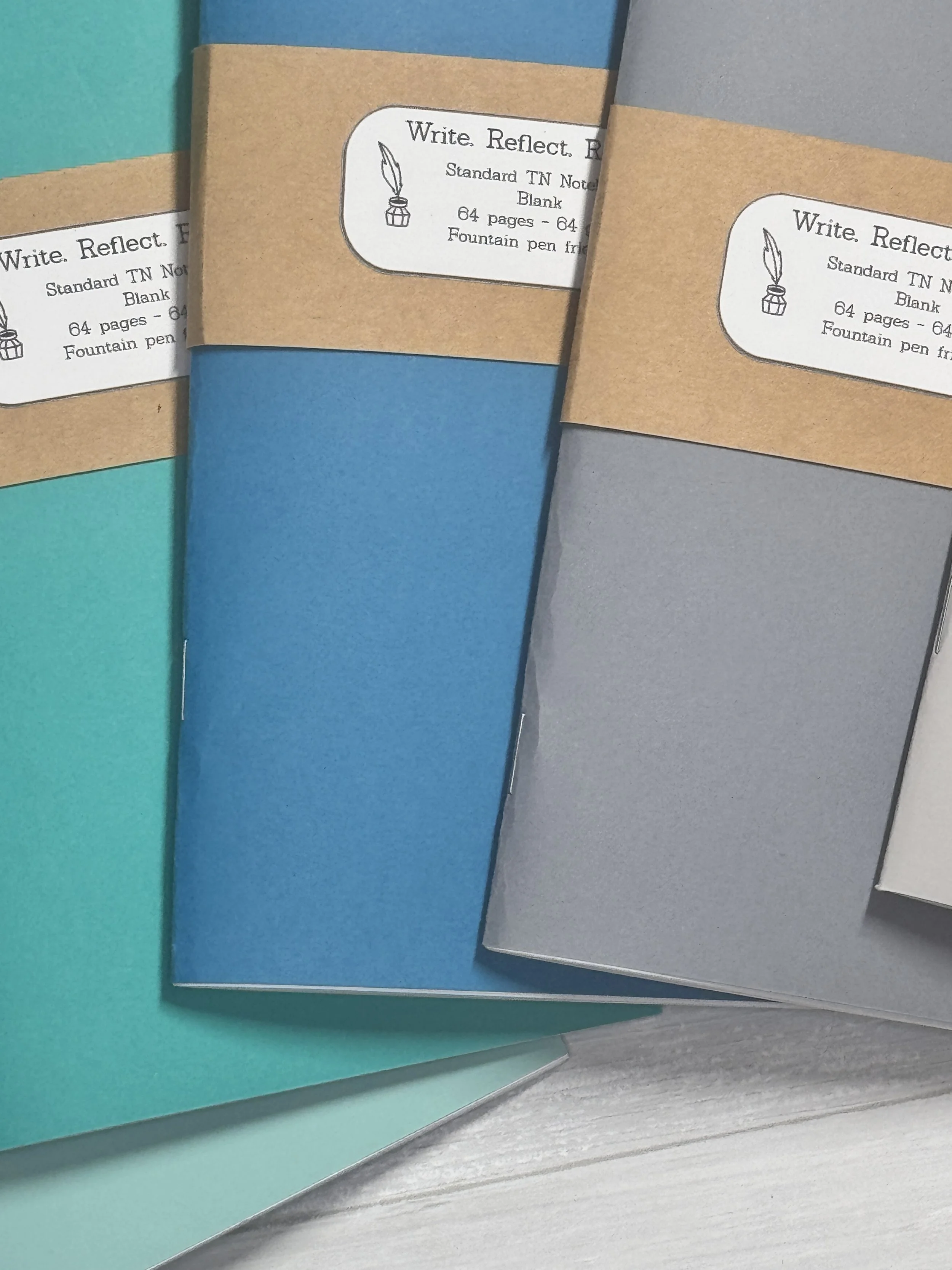 Multiple colored notebooks with kraft paper band on each, labeled as 'Write. Reflect. Review.' and 'Standard TN Notebook, Blank, 64 pages, fountain pen friendly.'