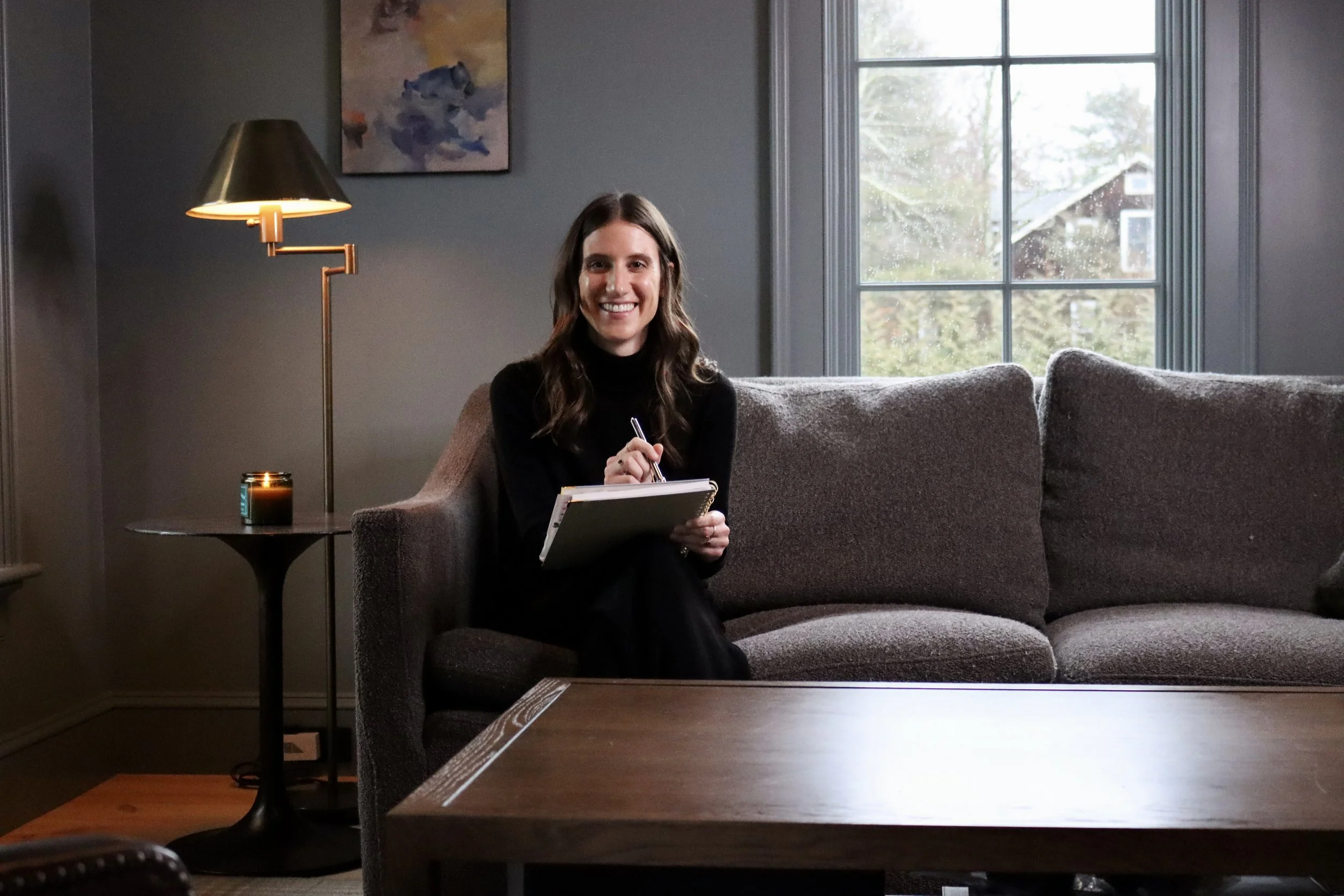 A woman with long brown hair, wearing a black turtleneck, sitting on a gray sofa in a living room, smiling while writing on a notepad. There is a window behind her, a floor lamp to her left, and a small table with a lit candle. An abstract painting hangs on the wall above the lamp.