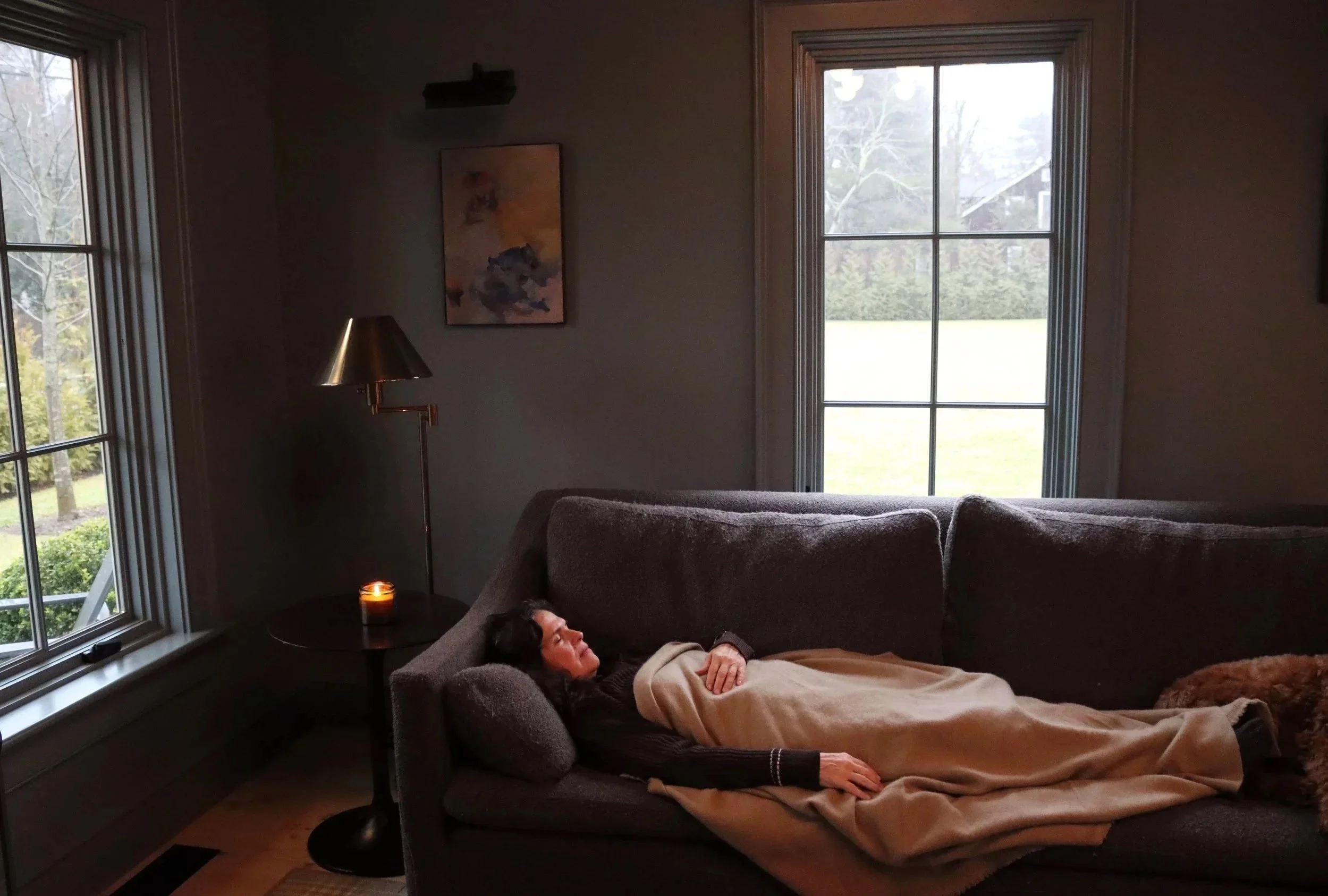 A woman resting on a dark gray couch in a living room, covered with a beige blanket, next to a window with a view of a grassy yard and trees outside. There is a lit candle on a small side table and a floor lamp nearby.