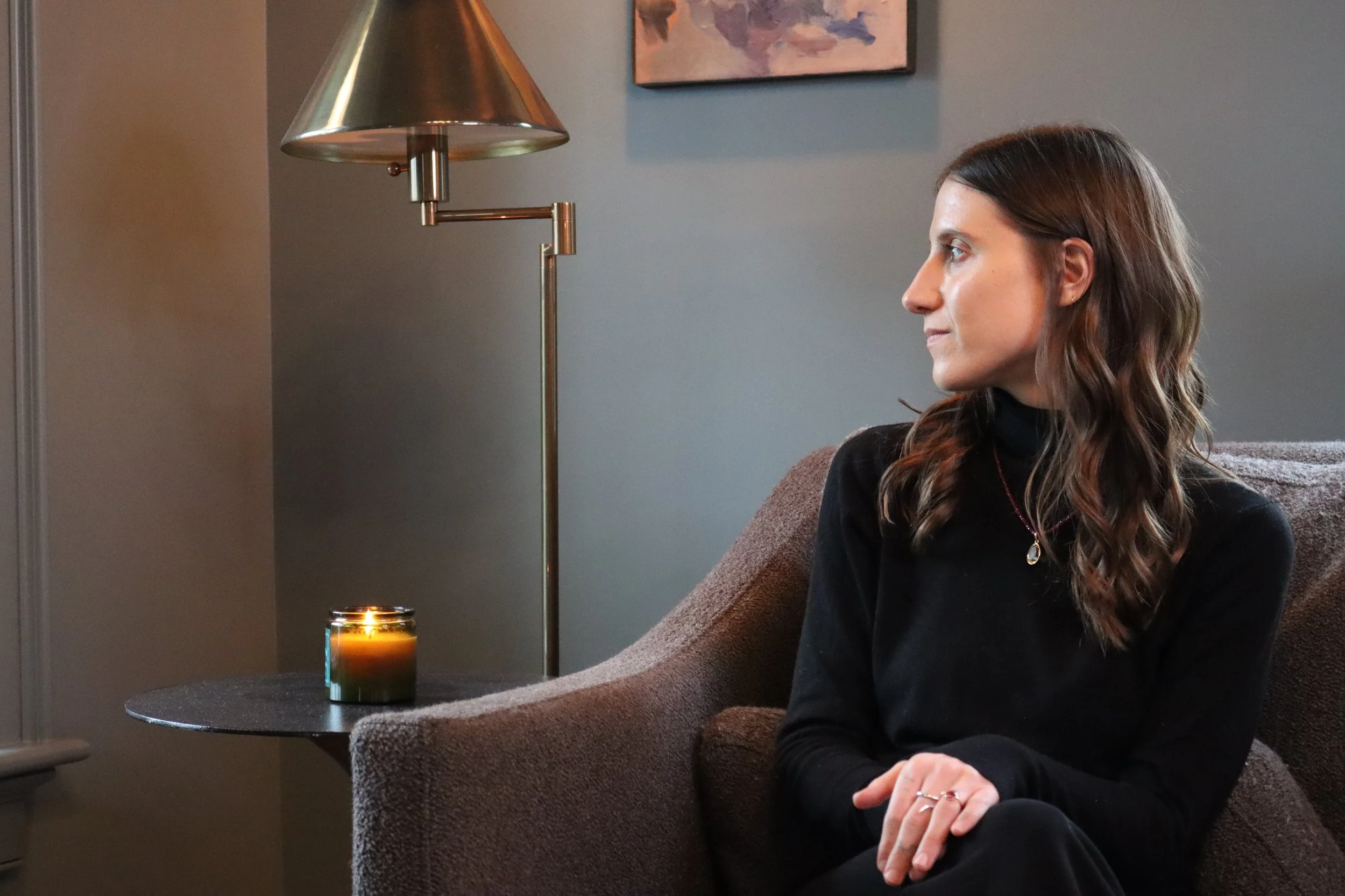 A woman with brown wavy hair wearing a black turtleneck sweater, sitting on a brown sofa, looking to her right with hands clasped. There is a small black side table with a lit candle, a brass floor lamp, and a wall painting in the background.