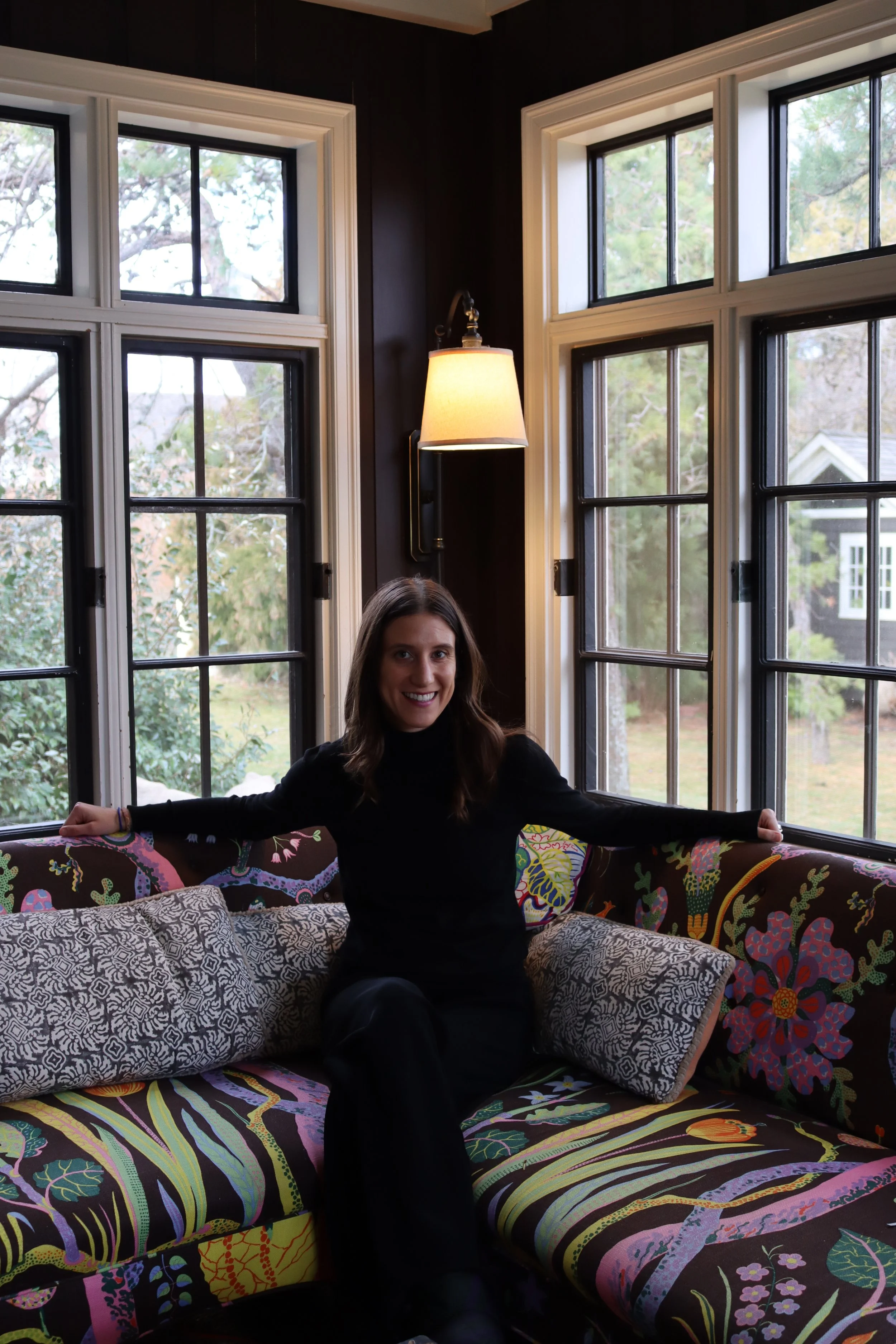 A woman in black clothing sitting on a colorful patterned sofa in front of large windows with a view of green trees outside, inside a cozy room with dark walls and a wall-mounted lamp.
