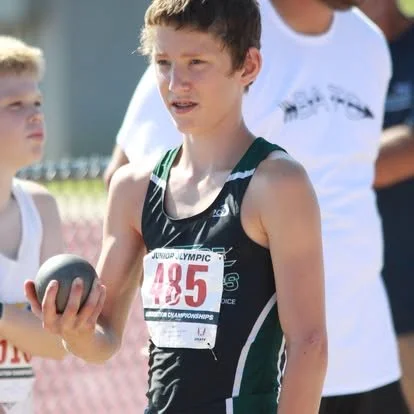 A teenage boy holding a shot put at an outdoor track and field event.