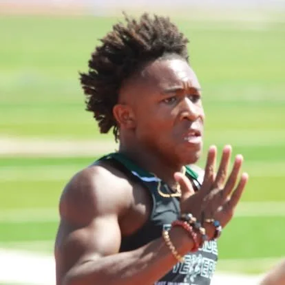 Close-up of a male runner with short curly hair, wearing a black athletic top, running outdoors on a track.