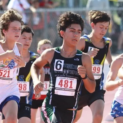 Young athletes competing in a race on a track field during daytime, wearing numbered bibs and running gear.