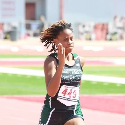 Young female runner with dreadlocks, wearing a black and green athletic uniform, running on a track field.