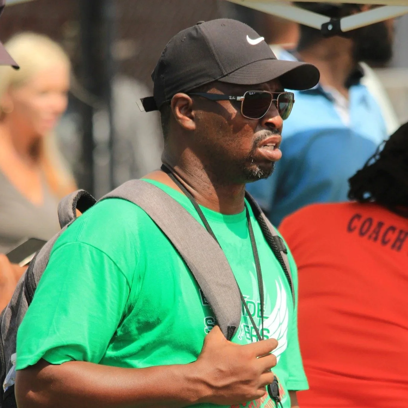Man in green shirt, black Nike cap, sunglasses, carrying a backpack at outdoor event.
