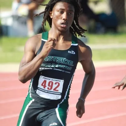 A female athlete running on a track during a race at the Junior Olympic event, wearing a black and green sports uniform with bib number 492.