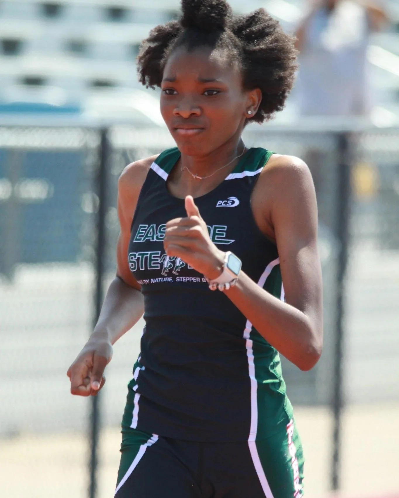 A female athlete with natural hair running outdoors on a track, wearing a black and green sports uniform.