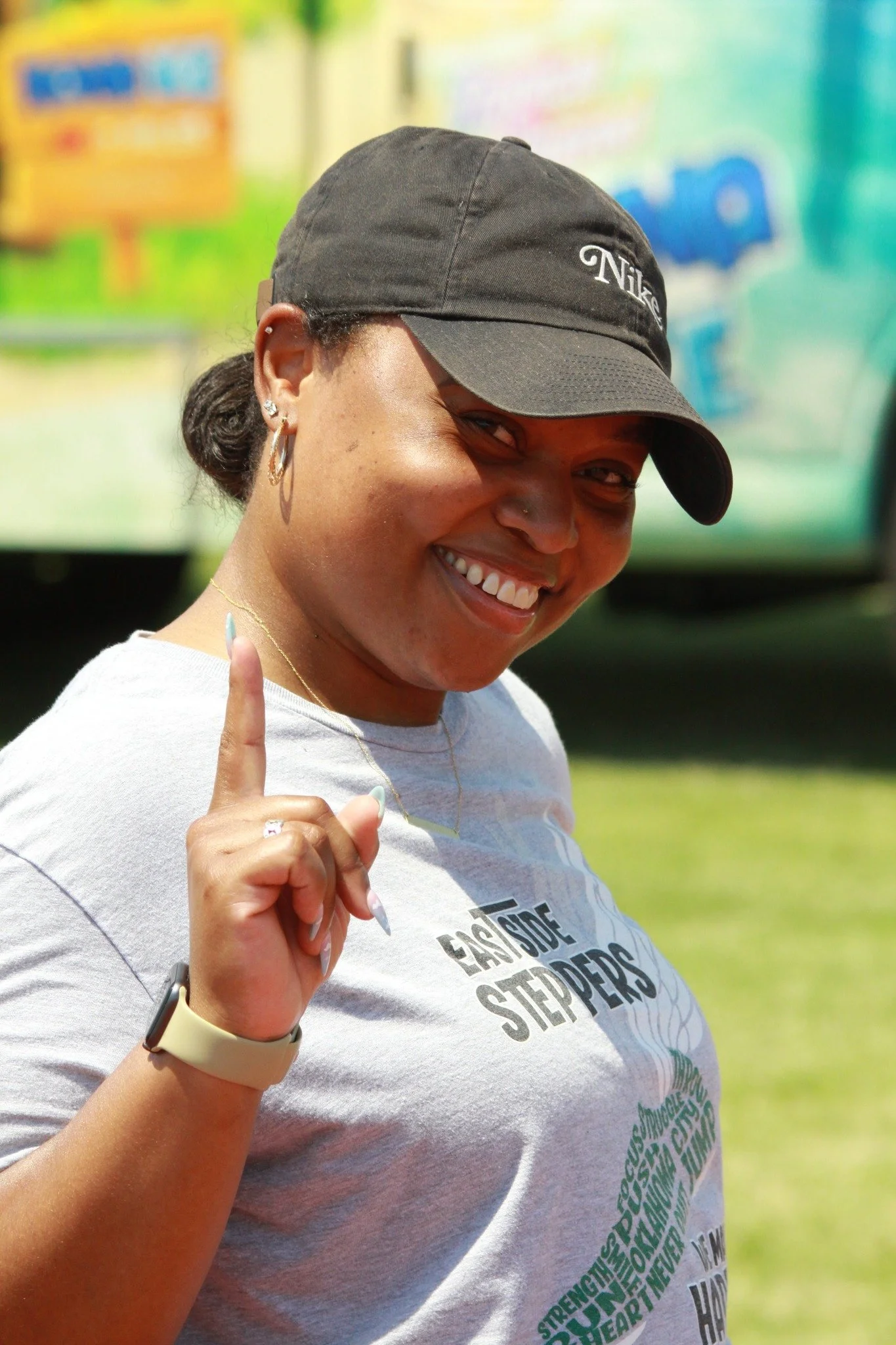 Smiling woman wearing a black Nike cap, hoop earrings, and a light gray T-shirt, holding up her index finger outdoors.