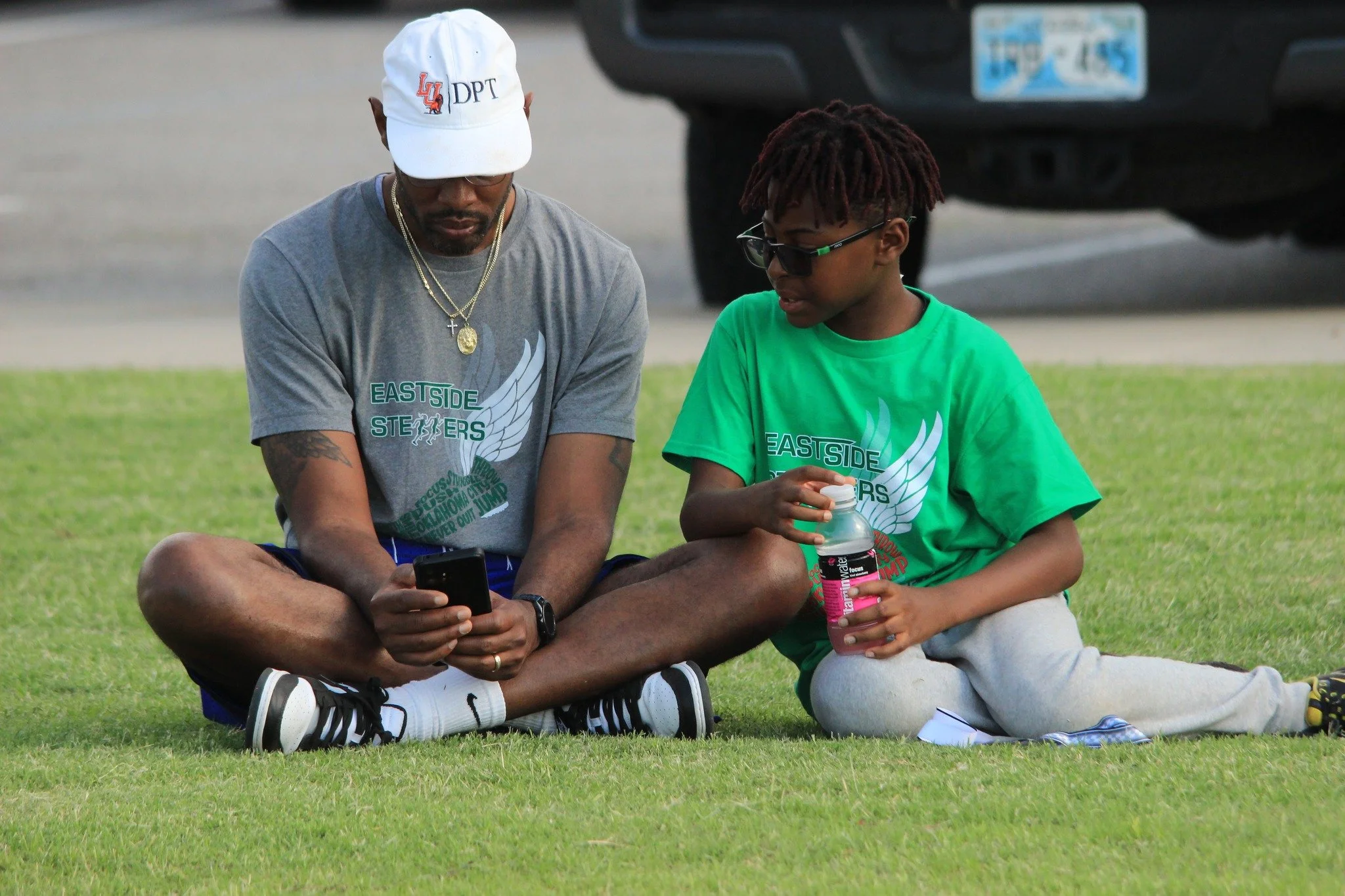 A man and a young boy sitting on grass, both wearing 'EASTSIDE STEPPERS' shirts. The man is looking at his phone, wearing a white cap, glasses, and jewelry. The boy is holding a bottle and wearing sunglasses.