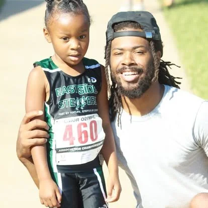 A man and a young girl standing outdoors. The girl is wearing a sports outfit with a race number bib, and the man is smiling with a cap backwards. They are likely participating in or celebrating a running event.
