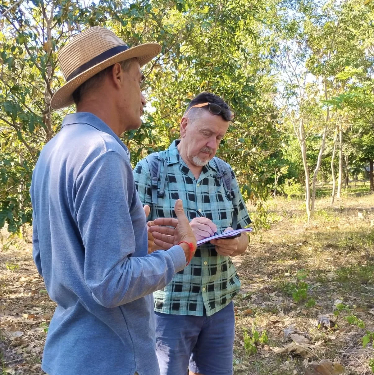 Two men standing outdoors in a wooded area, one wearing a straw hat and the other taking notes on a clipboard.