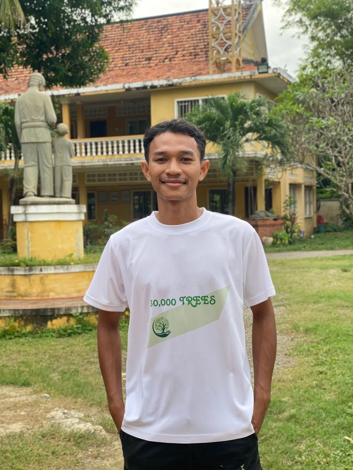 Young man in yellow shirt and camouflage hat holding a small potted tree outdoors, with more potted trees on a table and green foliage in the background.