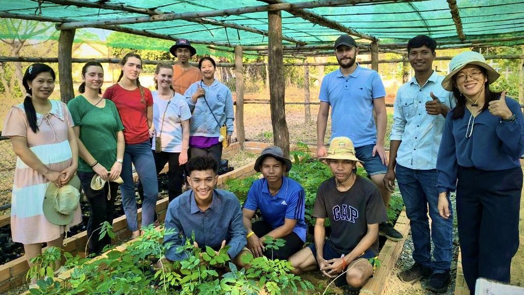 Group of eleven people, including men and women, standing and sitting in a garden or farm with young plants. Some are wearing hats and casual clothes, indicating an outdoor activity related to gardening or farming.