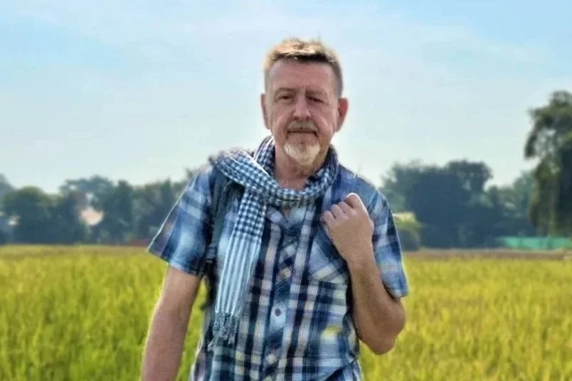 A man with a beard and short hair standing in a field, wearing a plaid shirt and a checkered scarf, with trees and a blue sky in the background.
