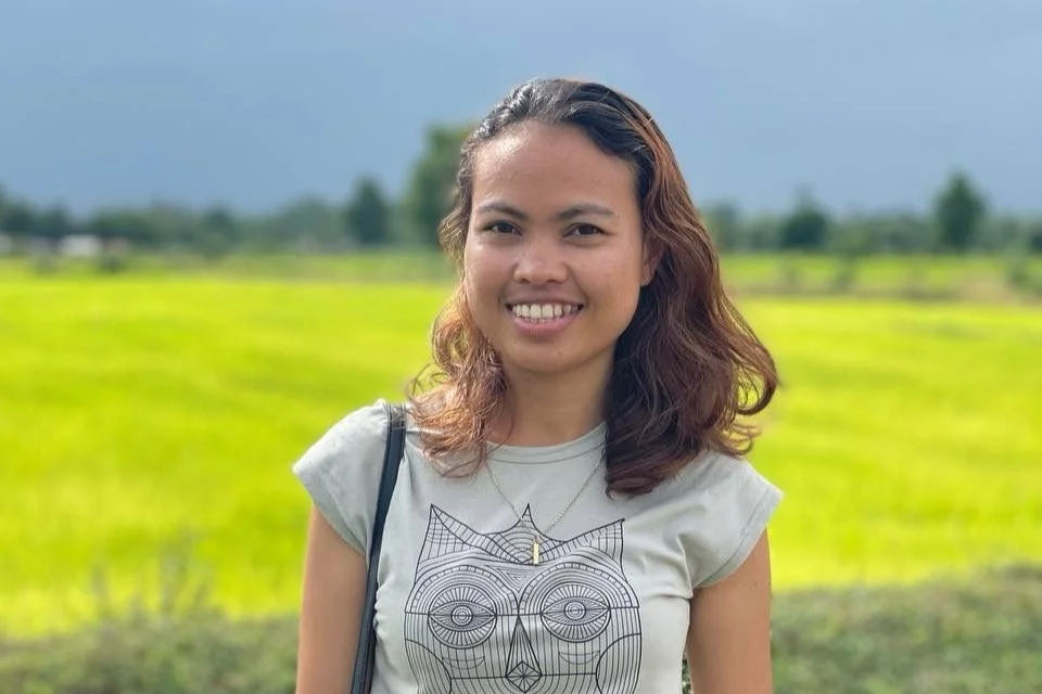A woman with shoulder-length wavy hair smiling outdoors in front of a green field under a cloudy sky, wearing a light gray t-shirt with a geometric owl design and a small necklace.