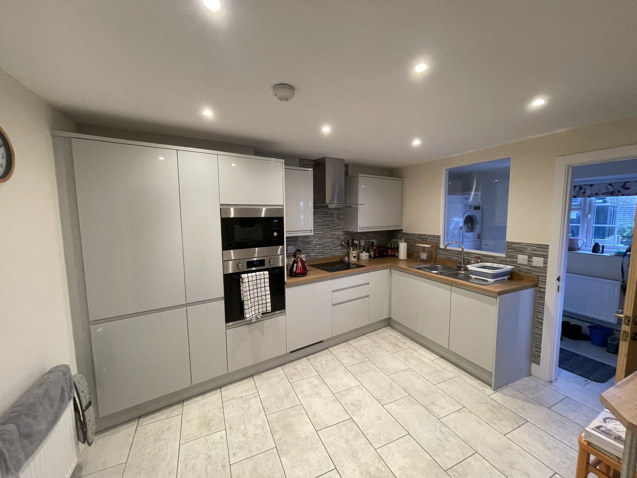 Modern kitchen with white cabinets, wooden countertops, gray backsplash, stainless steel appliances, and tiled floor. A doorway leads to a laundry area with a washing machine visible through the window.