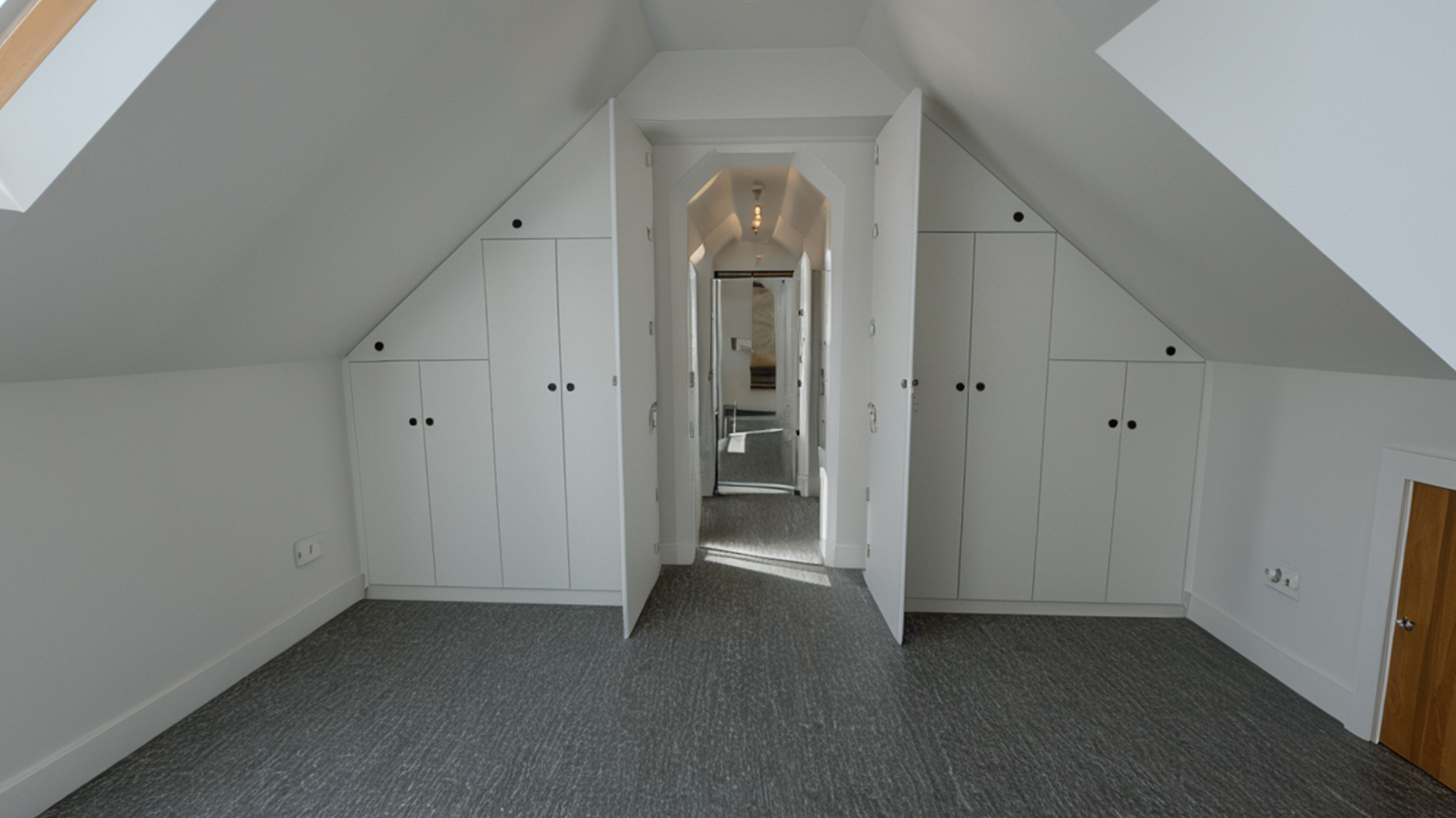 An attic bedroom with white built-in cabinets on both sides of a small door leading to a hallway, with sloped ceilings and a patterned gray carpet.