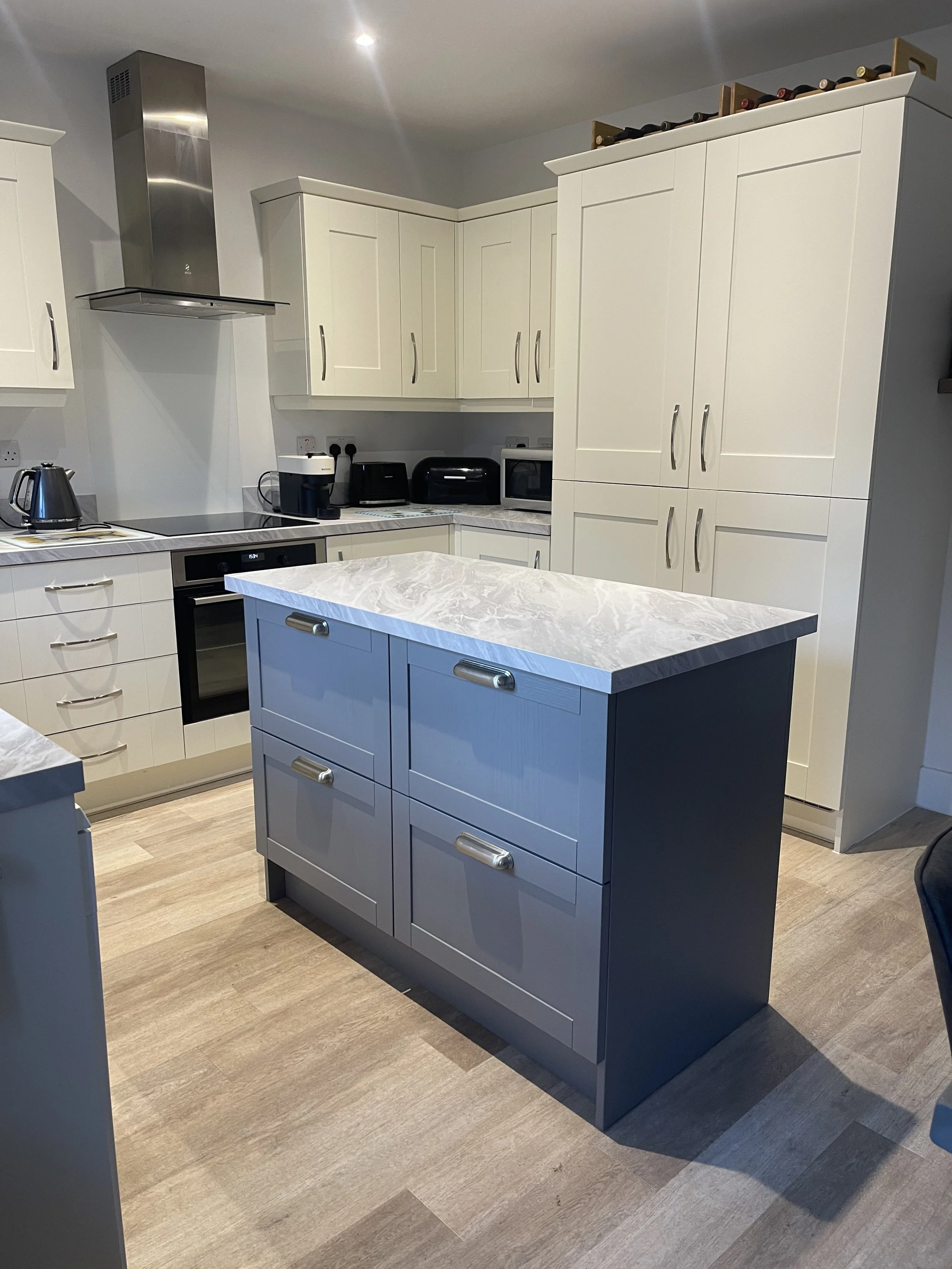 Modern kitchen with white cabinets, a blue kitchen island with drawers, marble countertop, and stainless steel appliances including a stove, microwave, and range hood. There is a wine rack on top of the cabinets.
