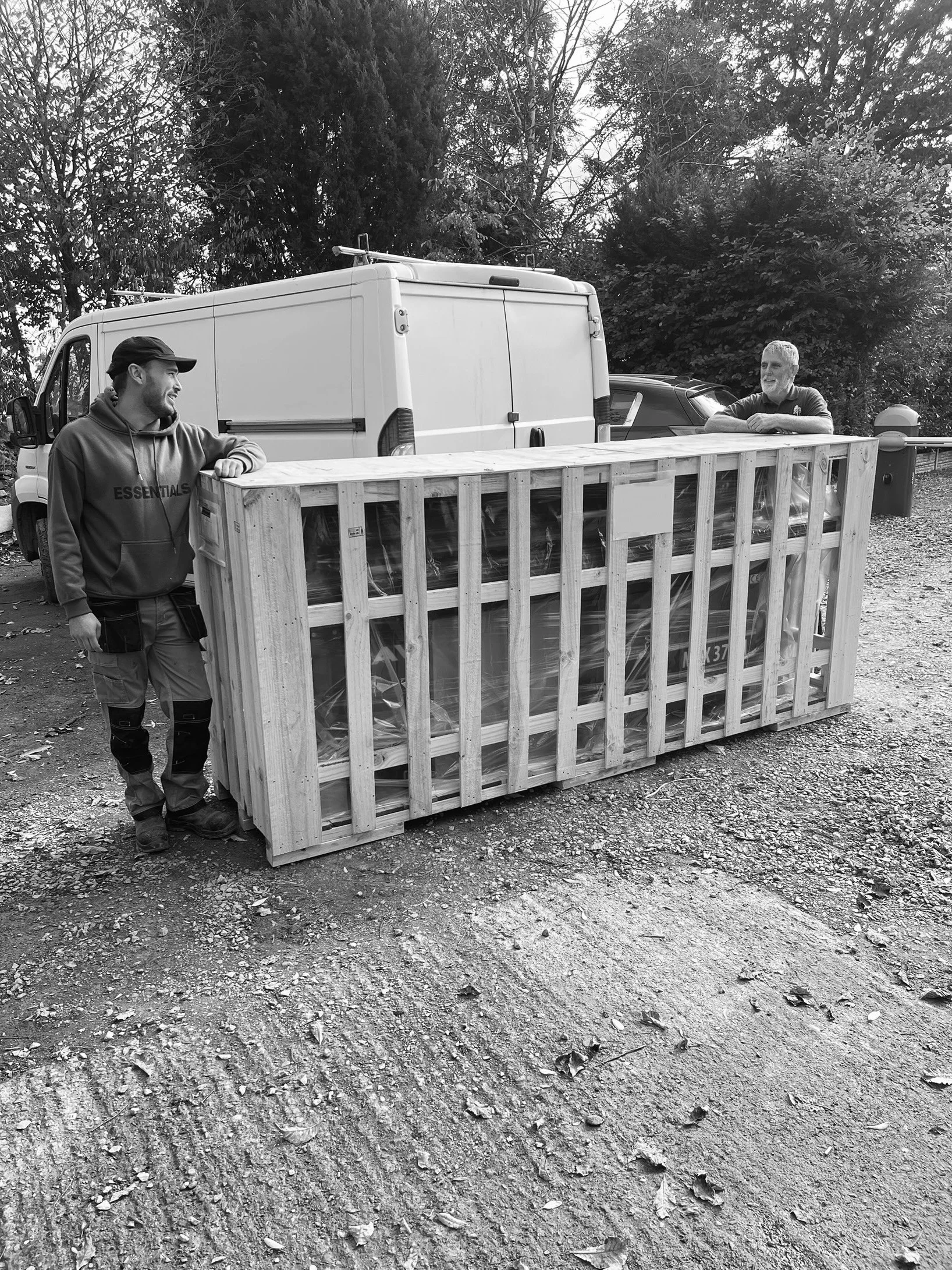 Two men in an outdoor setting with trees and parked vehicles, standing beside a large wooden crate with a transparent cover, leaning on or standing near it, and smiling at each other.