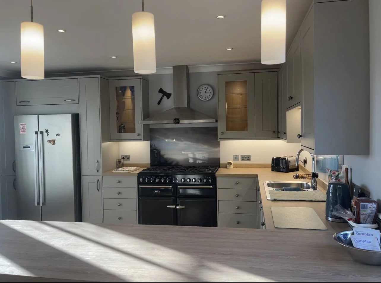 Kitchen with gray cabinets, black stove, stainless steel refrigerator, beige countertops, and various small appliances, illuminated by ceiling lights and hanging pendant lights.