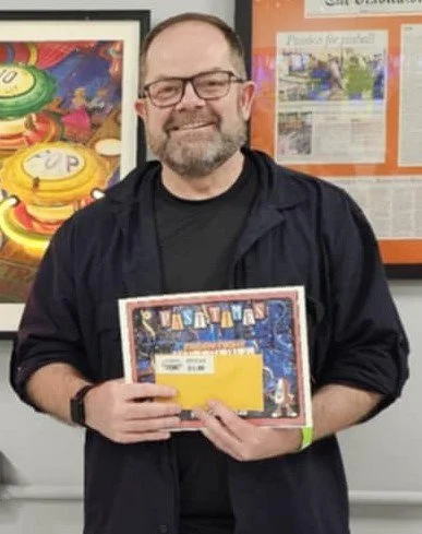 Man with glasses and beard smiling, holding a colorful puzzle box in a room with framed posters and artwork.