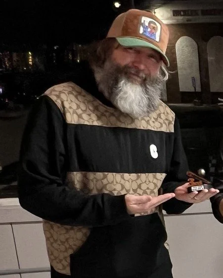 A smiling man with a gray beard, wearing a tan and black patterns hoodie and a baseball cap with a picture on the front, holding a small object in his hands.