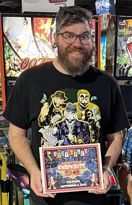 A man with glasses and a beard smiling, holding a colorful award certificate, standing in front of arcade game machines.