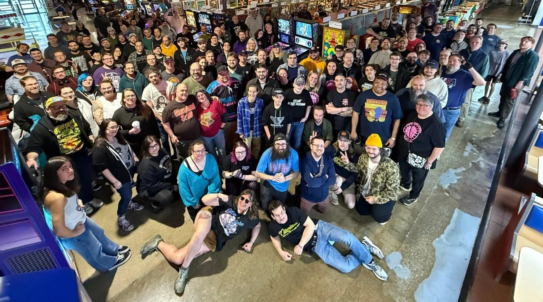 A large group of diverse people gathered in an arcade or entertainment area, looking up at the camera, with some standing and others kneeling or sitting in front. The background features arcade machines and colorful signage.