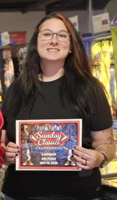 Woman with long dark hair, glasses, and tattoos holding a sign for a Sunday Classics Tournament at an arcade or gaming venue.