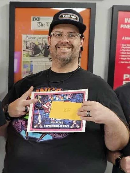 A man wearing glasses, a hat that says 'TOO HIP', and a black T-shirt, smiling and holding a colorful certificate or award with a yellow section, along with a pink-bordered document, while making a peace sign with his fingers.