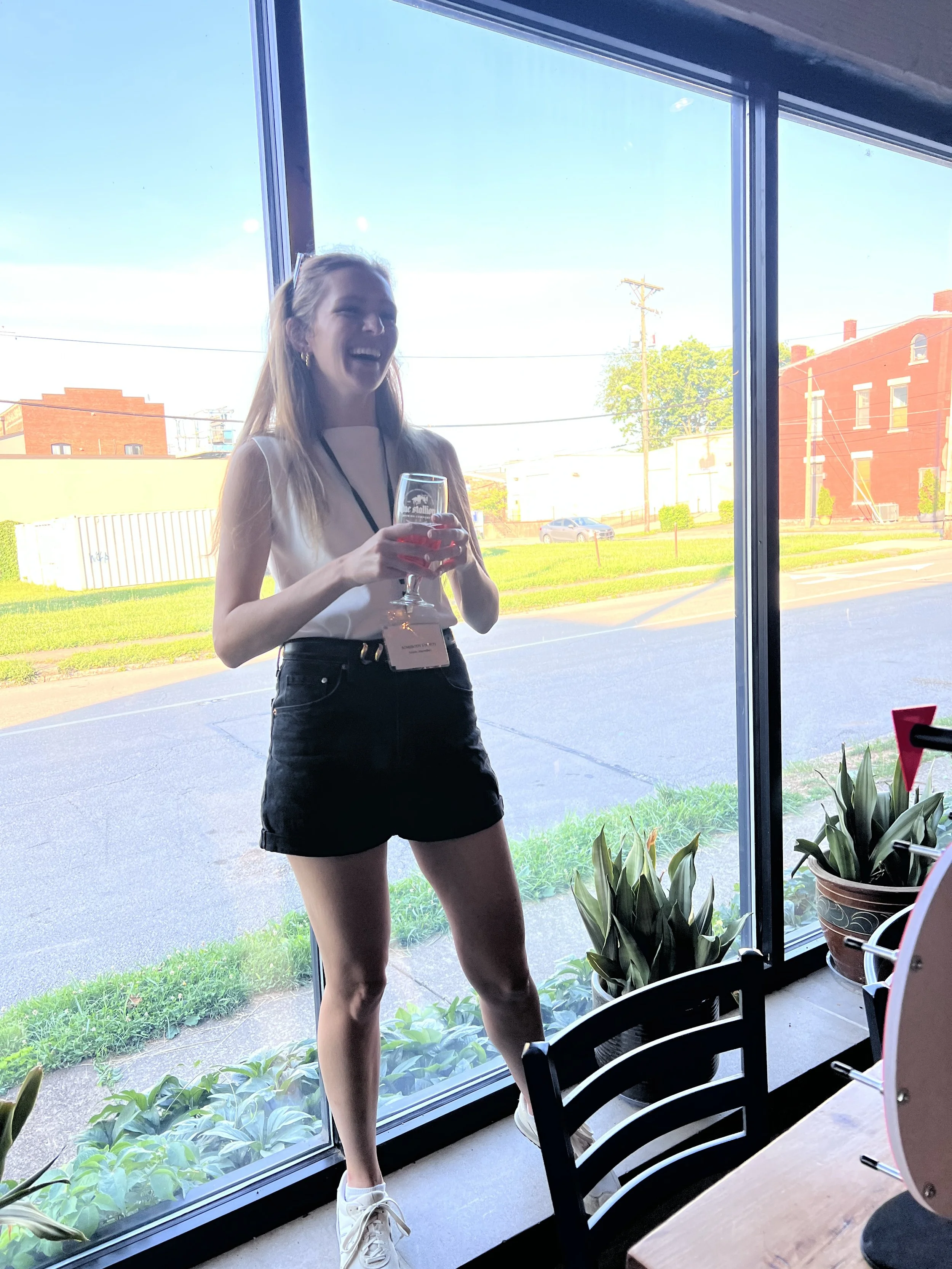 A young woman with long blonde hair, wearing a white sleeveless top, black shorts, and white sneakers, standing by a large glass window, holding a wine glass, smiling and laughing as she hosts a social club event.