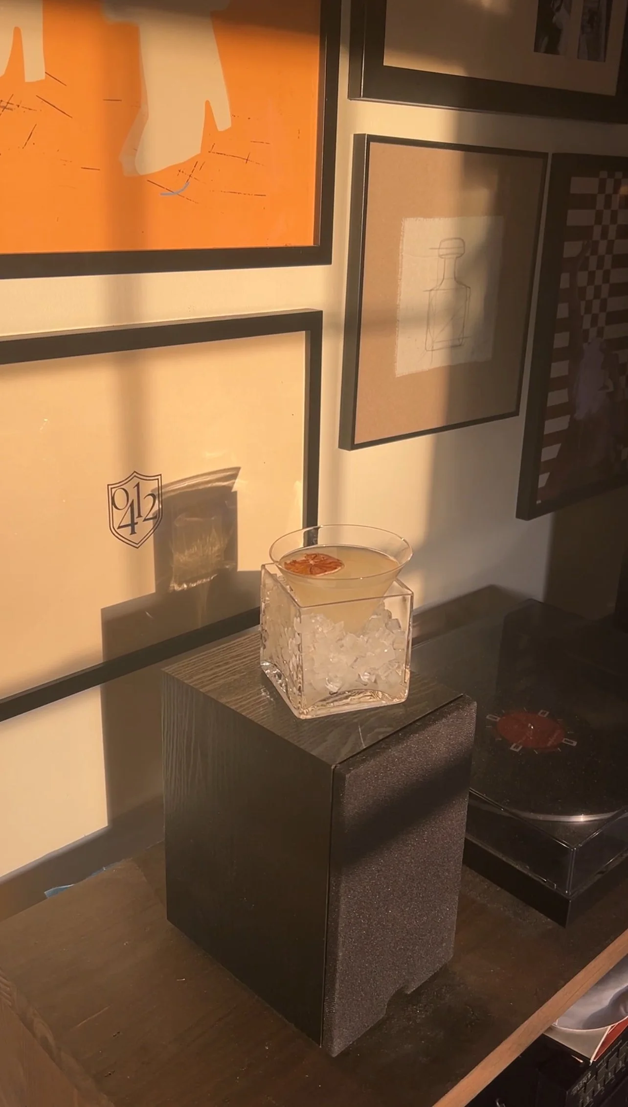 A vintage stemless martini glass with a cocktail with ice in a square glass container on a wooden table.  There is a turntable to the right during golden hour.
