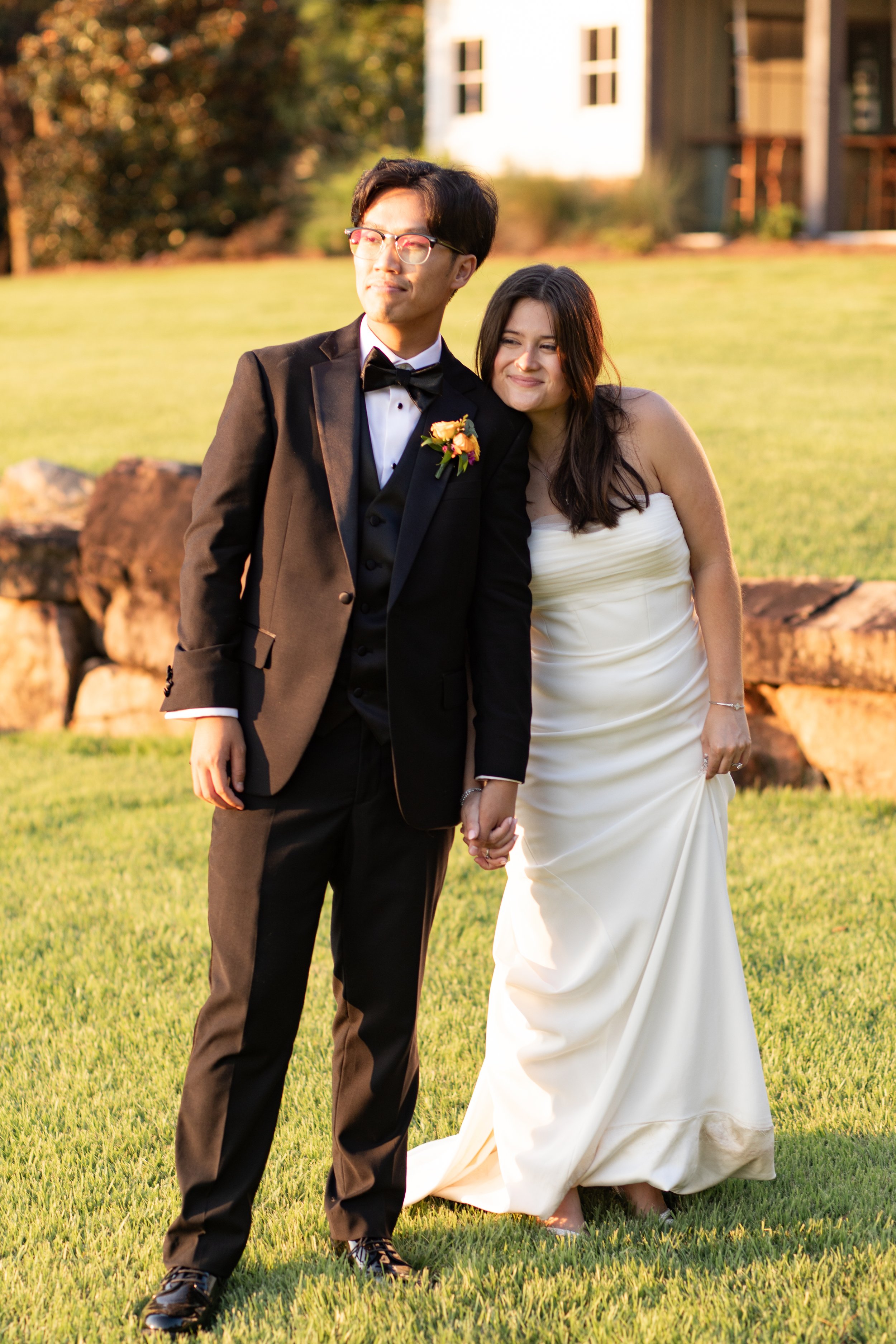 A newlywed couple holding hands outdoors during sunset, the groom wearing a black tuxedo with a boutonniere and glasses, the bride in a strapless white gown with long dark hair, both smiling softly.