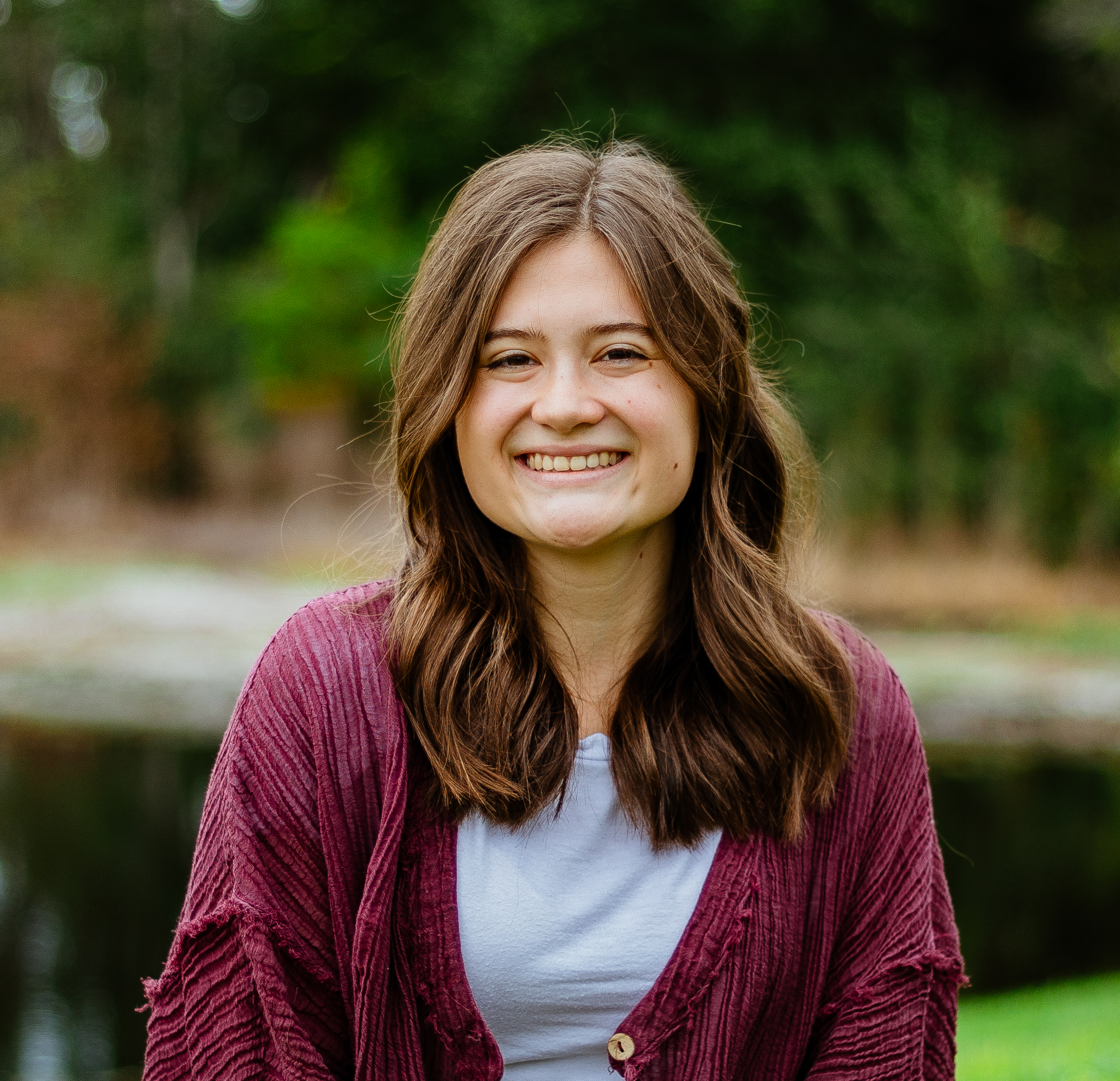 A smiling young woman with brown hair outdoors near a pond and trees.