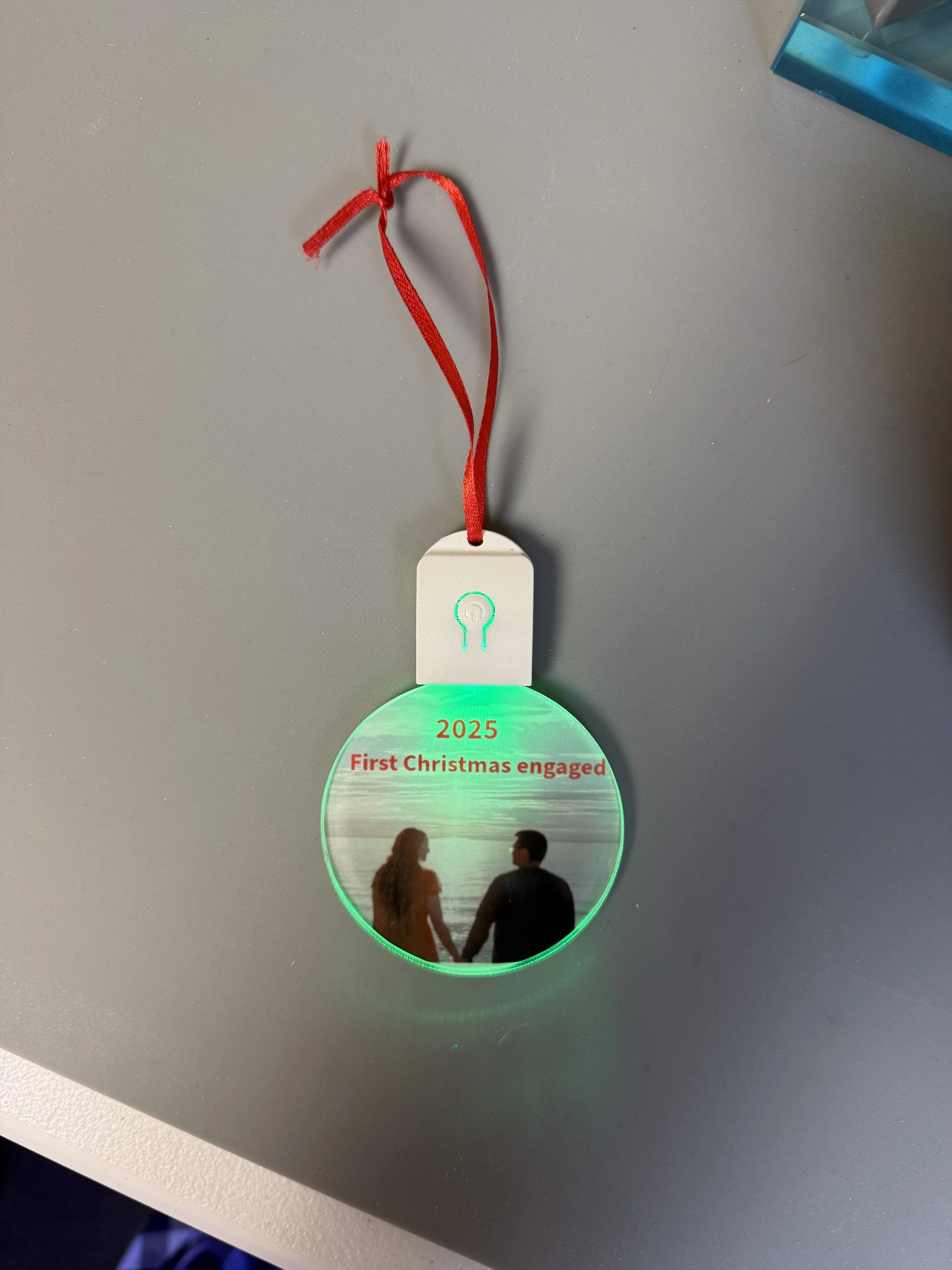 A round mirror with a photograph of a couple holding hands at the beach, illuminated with green light, with the text '2025 First Christmas engaged' above. The mirror is attached with a red ribbon to a white device on a gray surface.