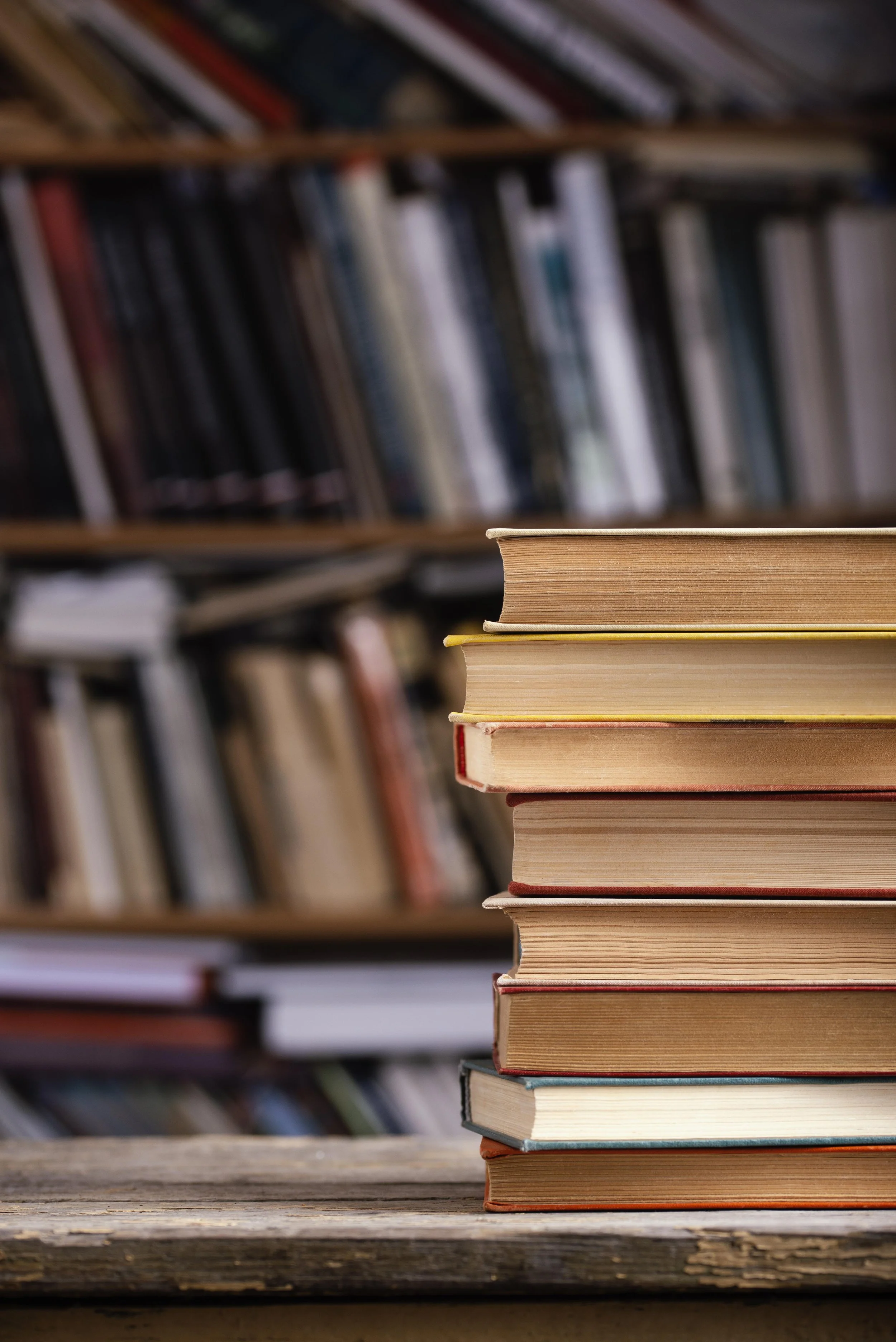 Stack of books on a wooden table with a bookshelf in the background.