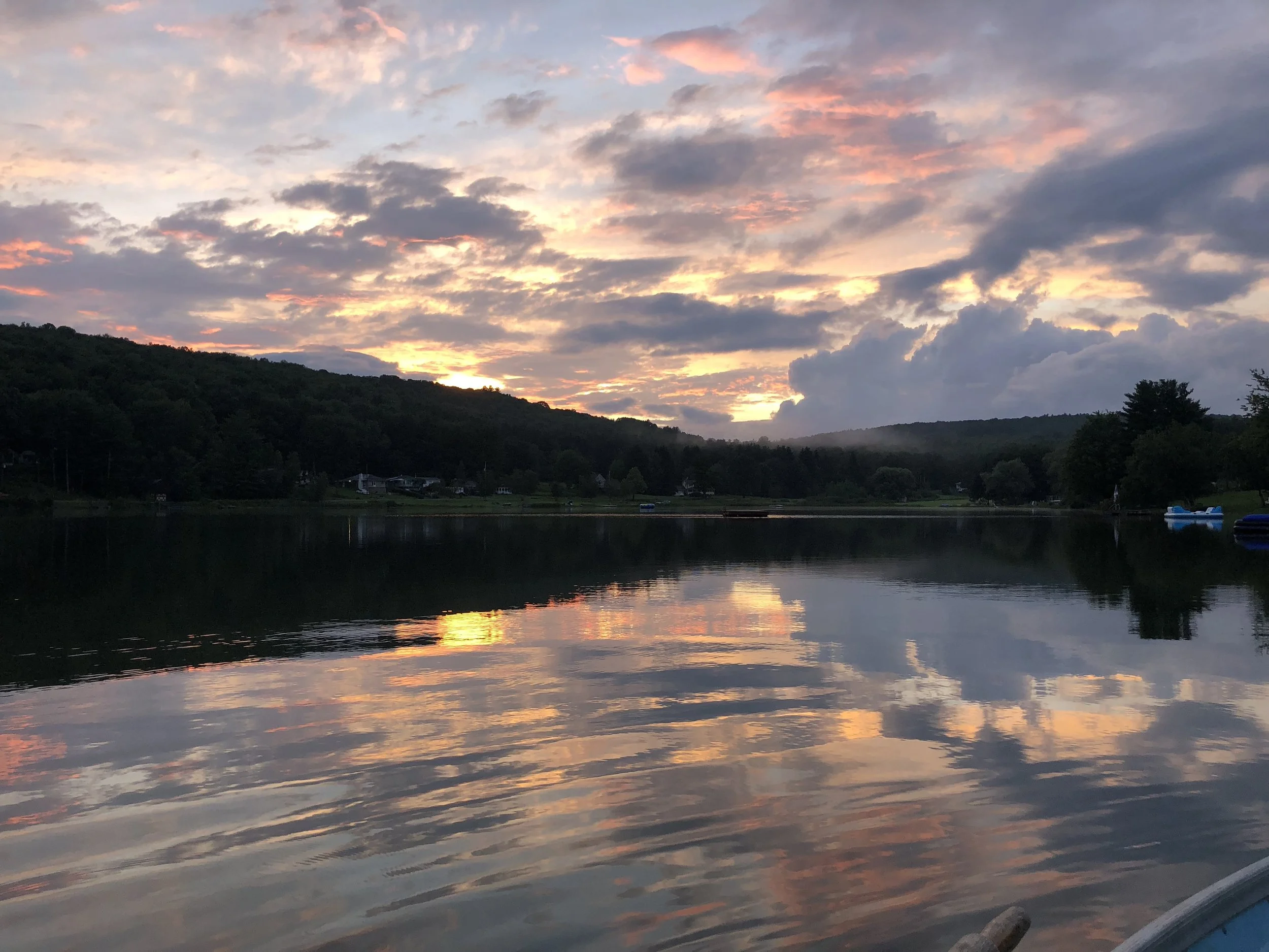 Sunset over a calm lake with reflections, surrounded by green hills and a few boats on the water.