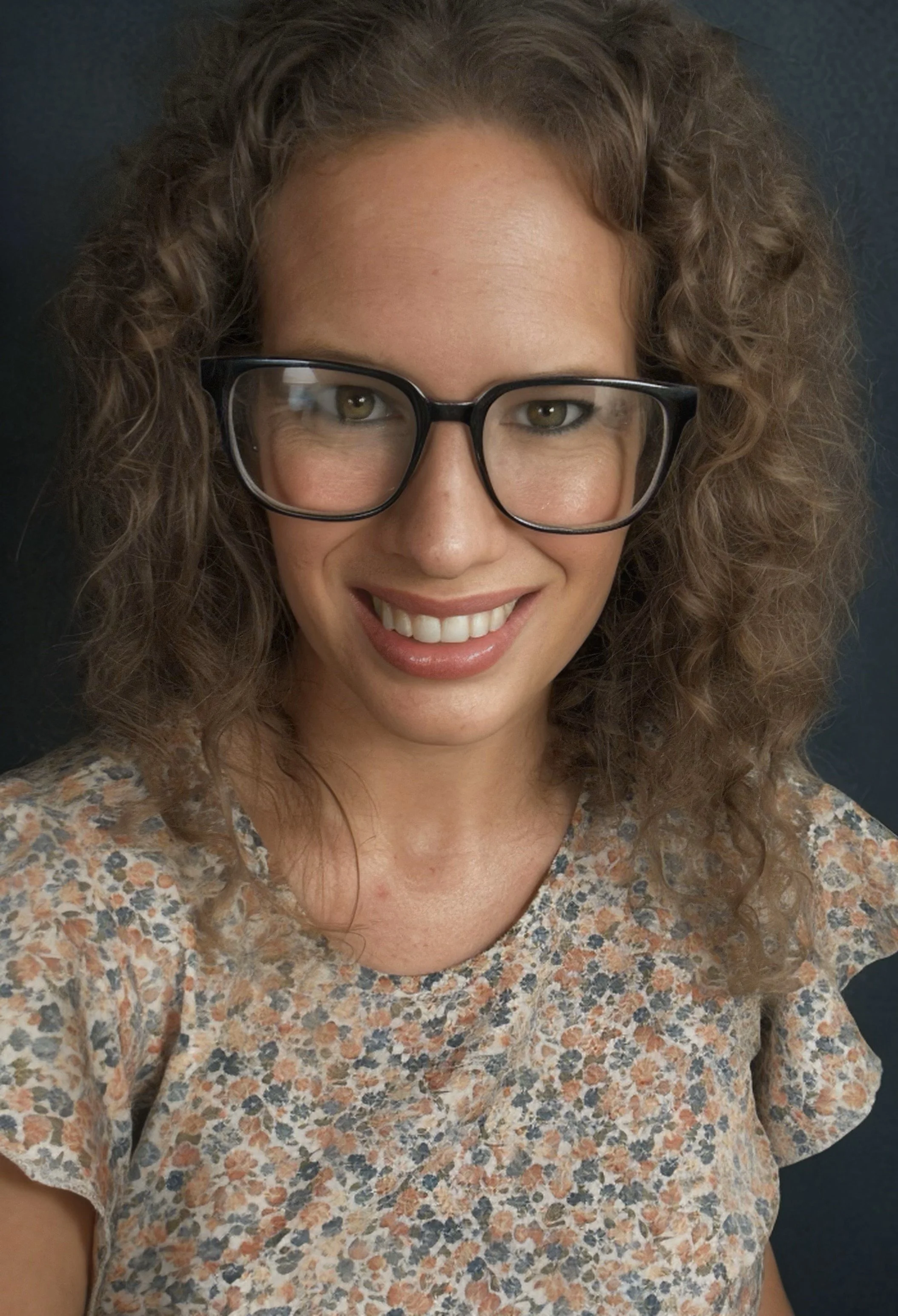 A woman with curly hair, wearing large black glasses and a floral patterned top, smiling at the camera.