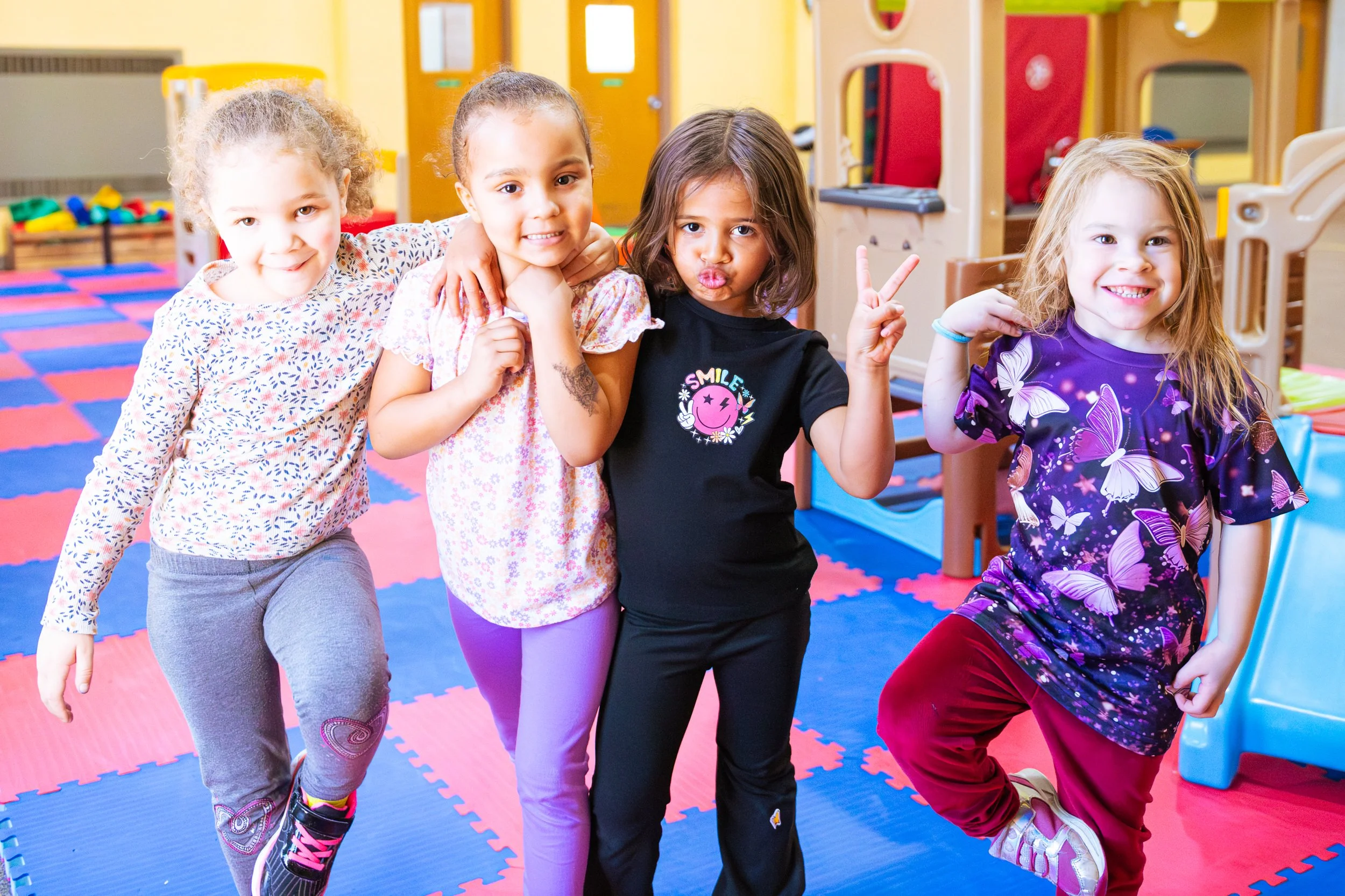 Four young girls standing together in a colorful indoor play area, smiling and making playful gestures.