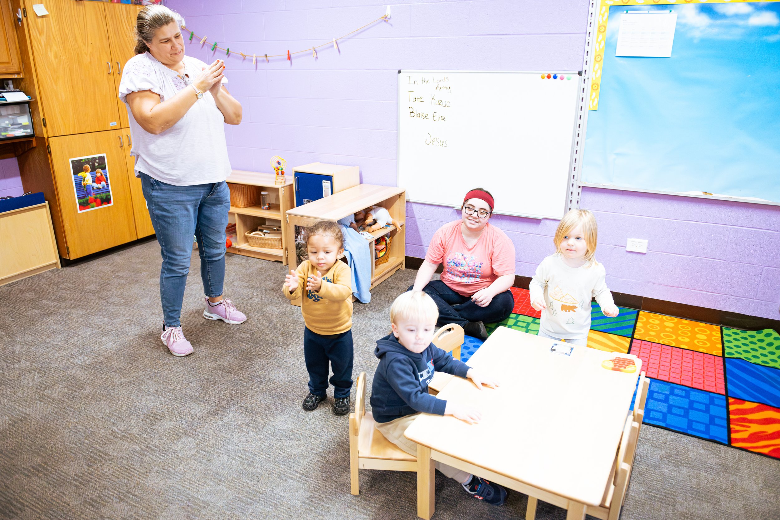 A preschool classroom with five children and two adults. One woman stands with clasped hands, while a girl and boy sit at a small table. Two additional children stand nearby, one with a yellow hoodie, and the other with a white shirt and blonde hair. A woman in a pink shirt and headband sits on the floor near the children.