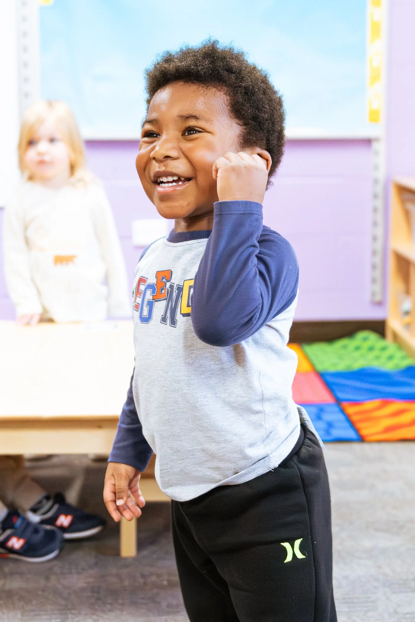 A young boy smiling and holding his hand to his ear inside a classroom, with a girl in the background.