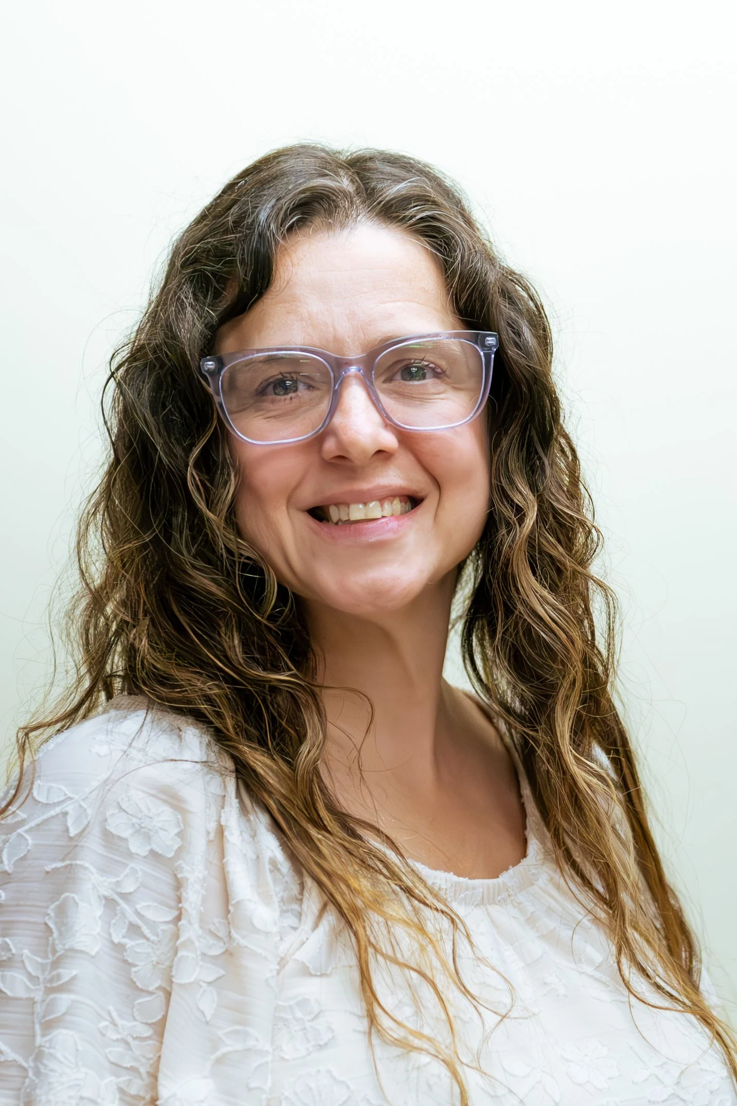 A woman with wavy brown hair, wearing clear glasses and a white embroidered blouse, smiling at the camera against a plain background.