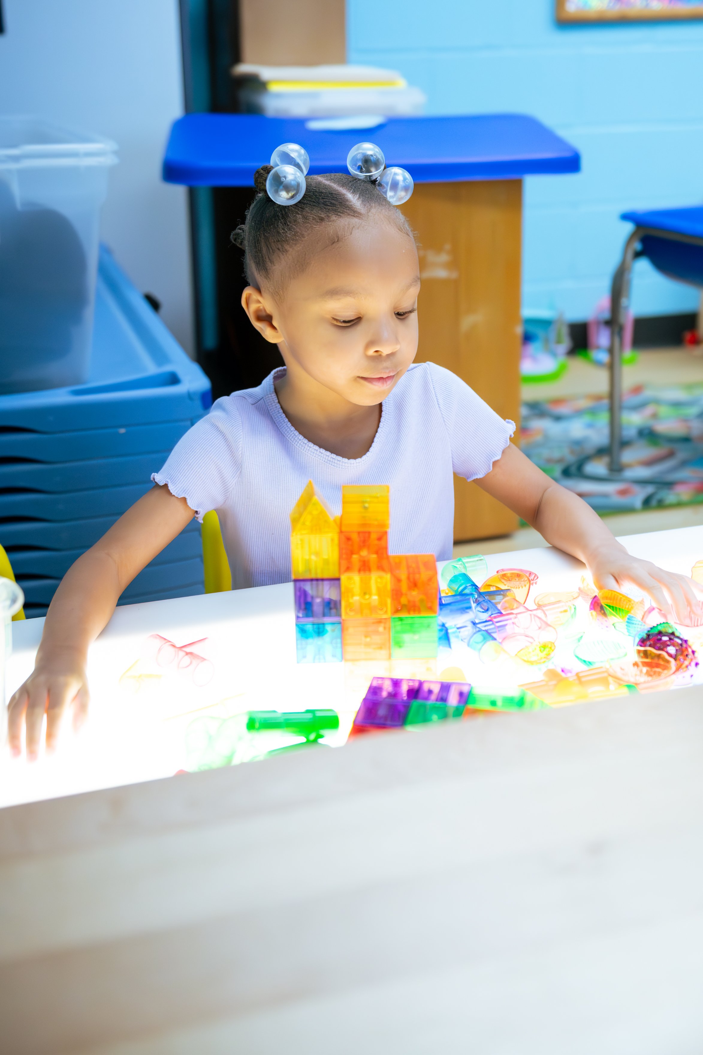 Young girl sitting at a table playing with illuminated colorful magnetic tiles, in a classroom setting.