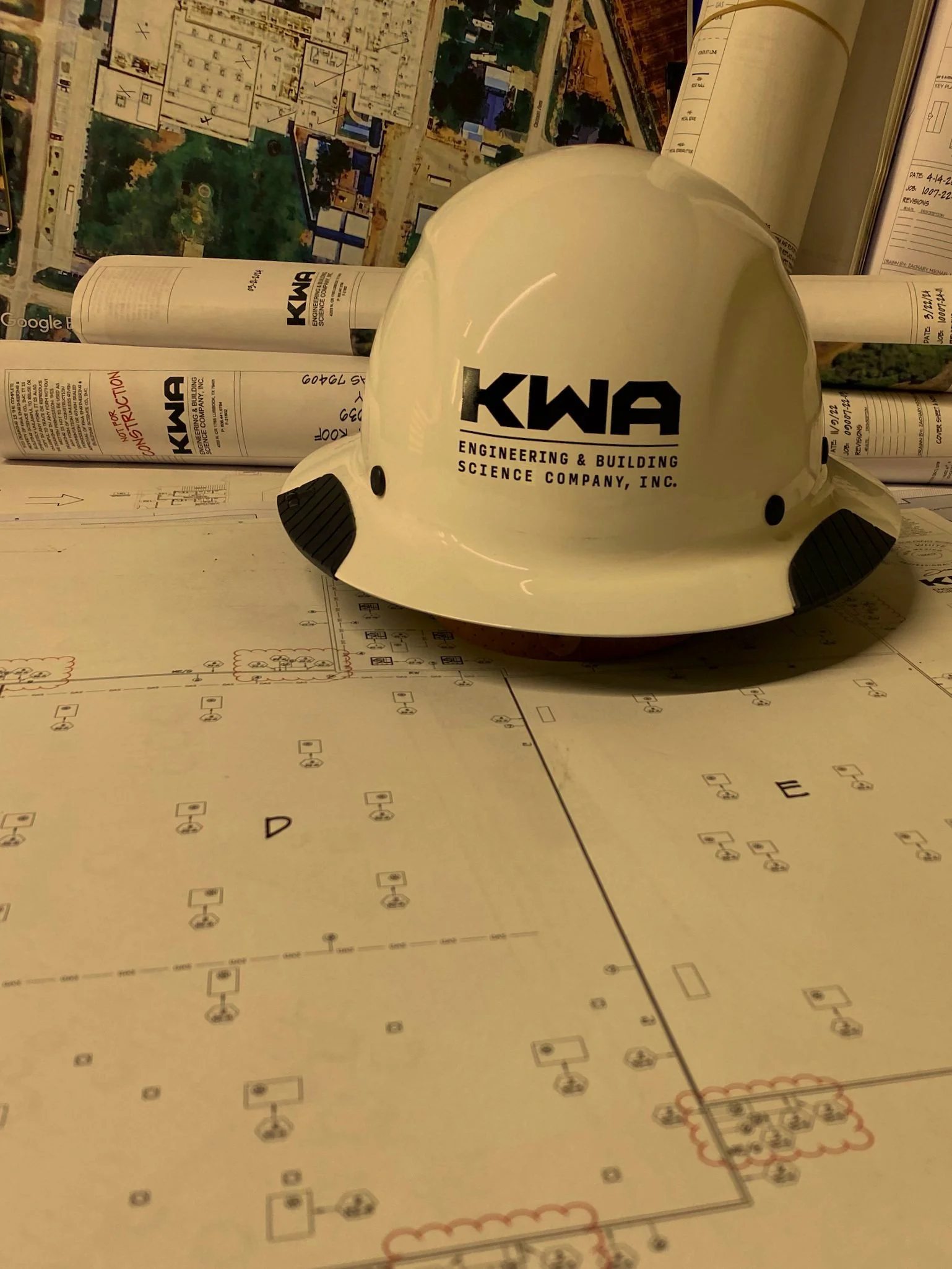 A white safety helmet with the company logo 'KWA' resting on technical blueprints, with rolled-up plans and a colorful map or aerial photograph in the background.