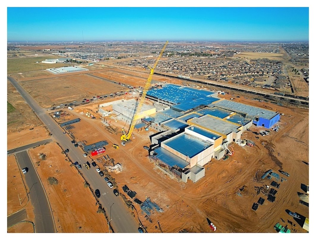 Aerial view of a large construction site for a new building or facility, with a yellow crane, construction vehicles, and partially completed structures, set in an area with dirt and roads, with a parking lot and a residential neighborhood in the background.